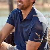 Man in a Coastal Polo Shirt by Tom Beckbe, sitting outdoors and smiling against a blurred natural background.