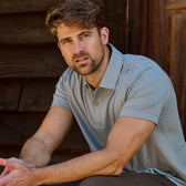 A brown-haired, bearded man sits by a wooden structure wearing a Tom Beckbe Coastal Polo Shirt.
