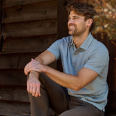 Person in a Coastal Polo Shirt by Tom Beckbe and dark pants, sitting outside near a wooden wall, smiling to the right.