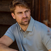 A man in a light gray Coastal Polo Shirt by Tom Beckbe sits outdoors, looking to the side with a neutral expression.
