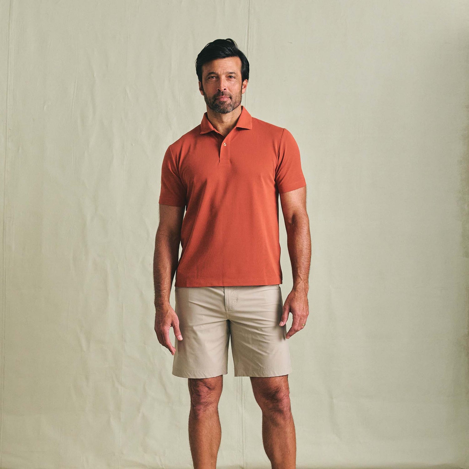 A man wears a rust Coastal Polo Shirt by Tom Beckbe and beige shorts against a plain background.