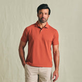 Man wears a Tom Beckbe Coastal Polo Shirt in orange with beige pants, standing against a plain light background.