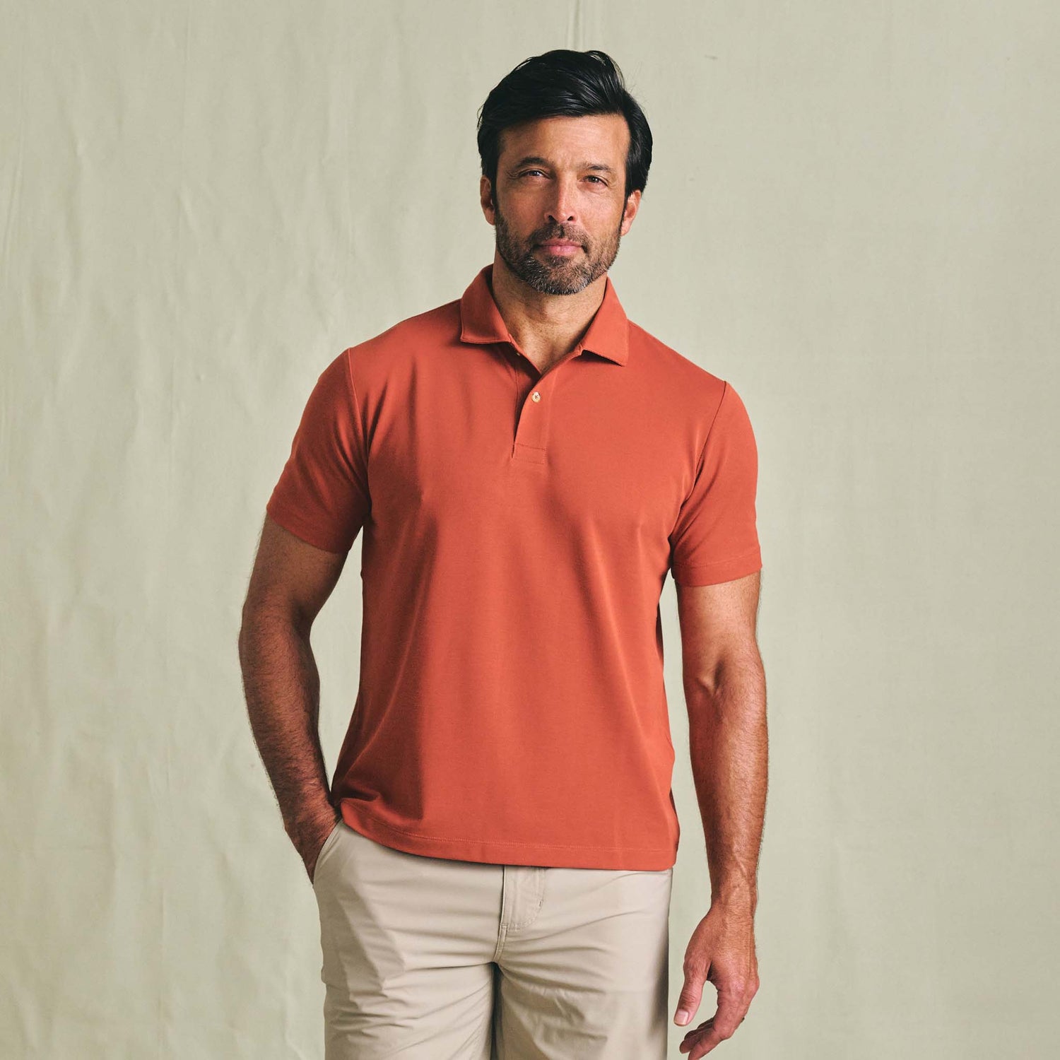 Man wears a Tom Beckbe Coastal Polo Shirt in orange with beige pants, standing against a plain light background.