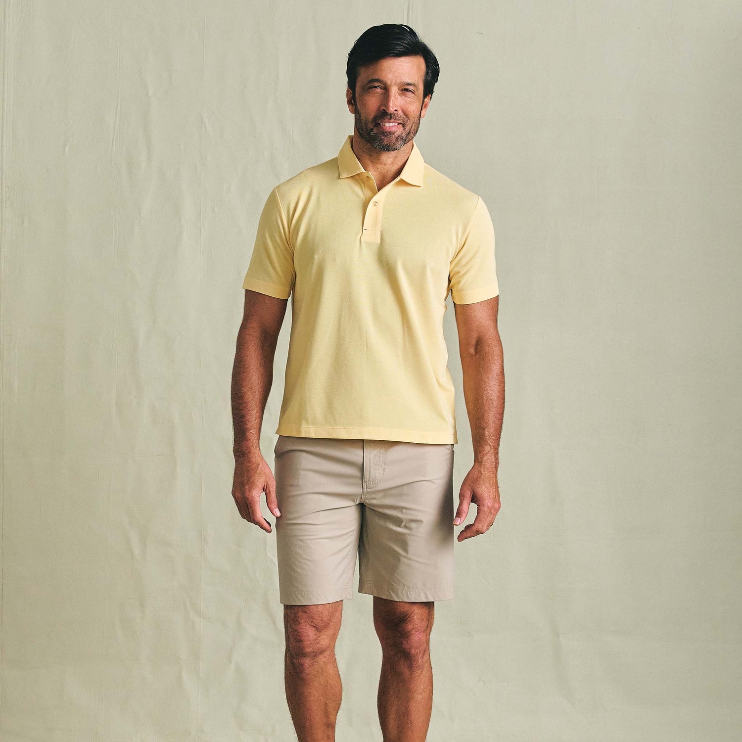 Man in a light yellow Tom Beckbe Coastal Polo Shirt and beige shorts stands against a light background.