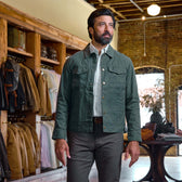 A man in a green Tom Beckbe Crawford Jacket and brown pants stands in a clothing store with shirts and jackets behind him.