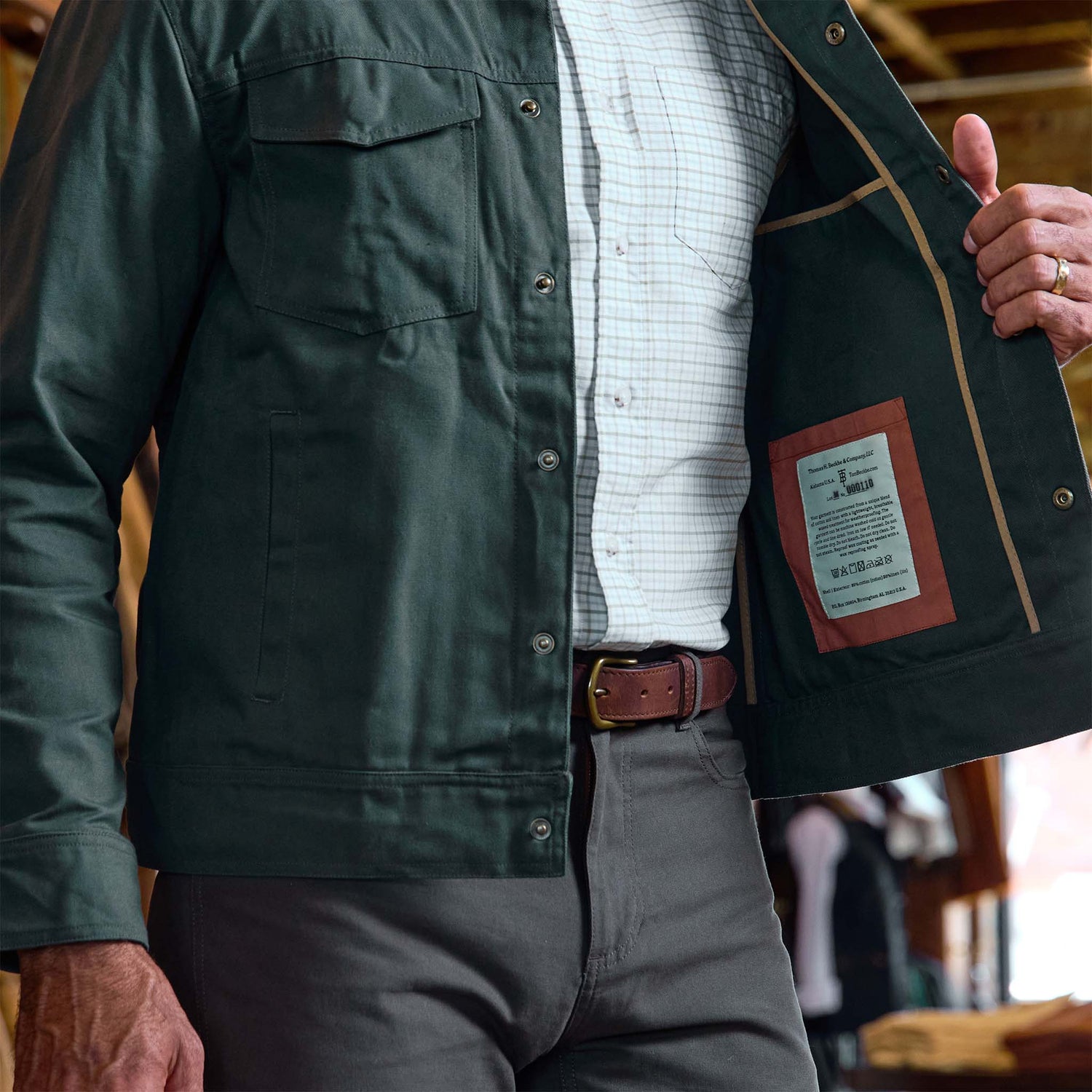 Man wearing the Tom Beckbe Crawford Jacket over a checked shirt, holding it open to display its interior label.