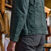 A man wearing the Tom Beckbe Crawford Jacket and gray pants stands indoors with clothing racks behind him.