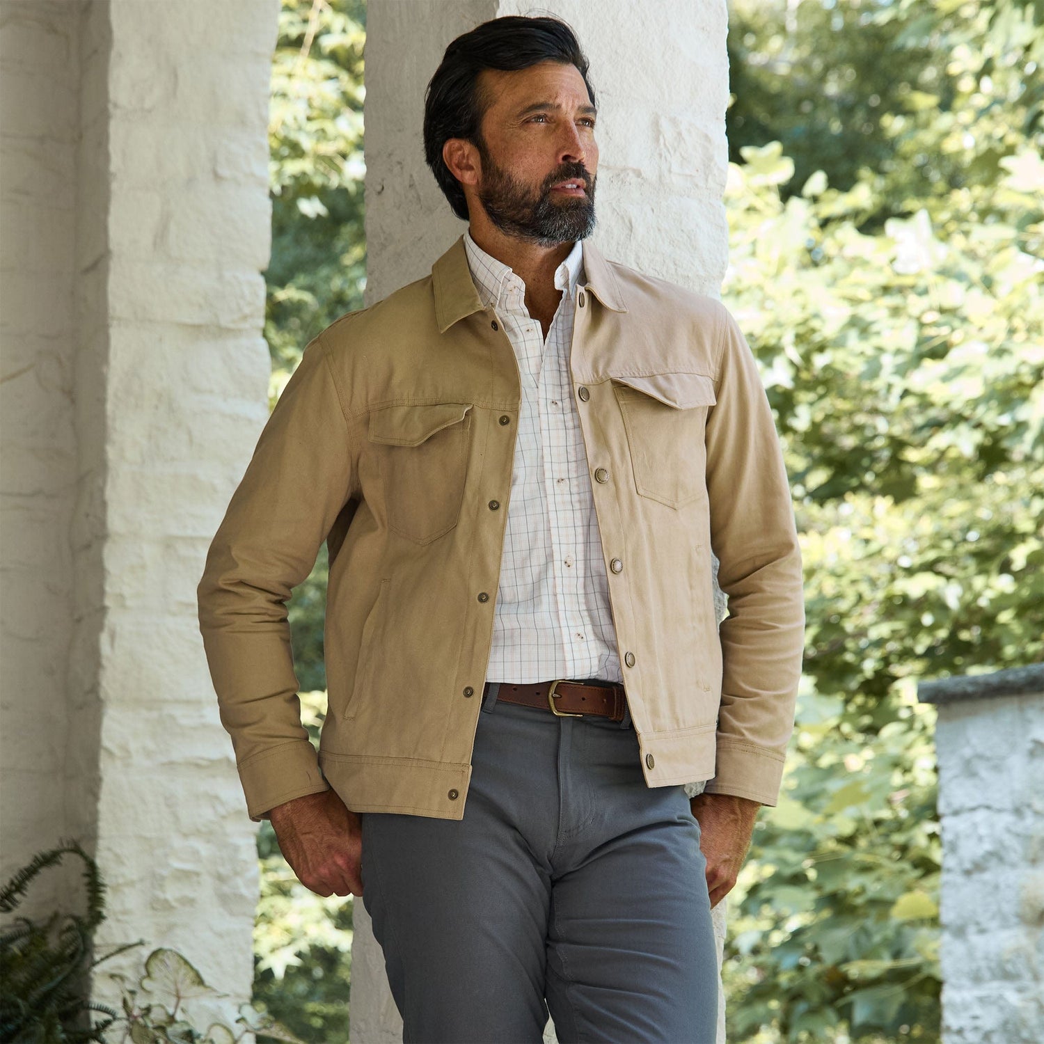 A man in a Tom Beckbe Crawford Jacket, plaid shirt, and gray pants stands by a white stone column outdoors.