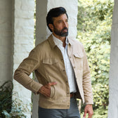 Man wearing Tom Beckbe’s Crawford Jacket over a plaid shirt stands on a porch with white pillars and greenery.