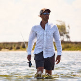 A man in Tom Beckbes Crenshaw Performance Shorts wades through waist-deep water, fishing under a partly cloudy sky.