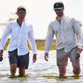 Two men in Tom Beckbe Crenshaw Performance Shorts, plaid shirts, and sunglasses walk through shallow water with fishing gear.