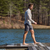A man in Crenshaw Performance Shorts by Tom Beckbe strolls barefoot on a dock by the lake, holding a fishing rod.