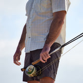Man in Crenshaw Performance Short and shirt by Tom Beckbe, holding a fishing rod against a pale sky backdrop.