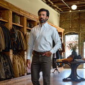 Man wears a Tom Beckbe Culver Oxford Shirt and dark pants in a clothing store with shirts and jackets behind him.