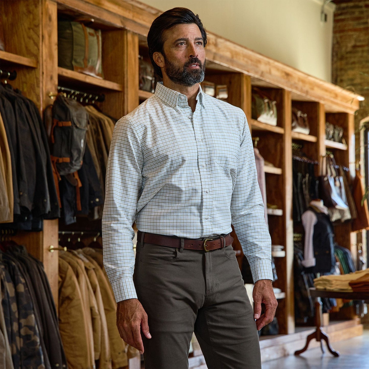 A man in a Tom Beckbe Culver Oxford Shirt and khaki pants stands in a store with wooden shelves and hanging jackets.