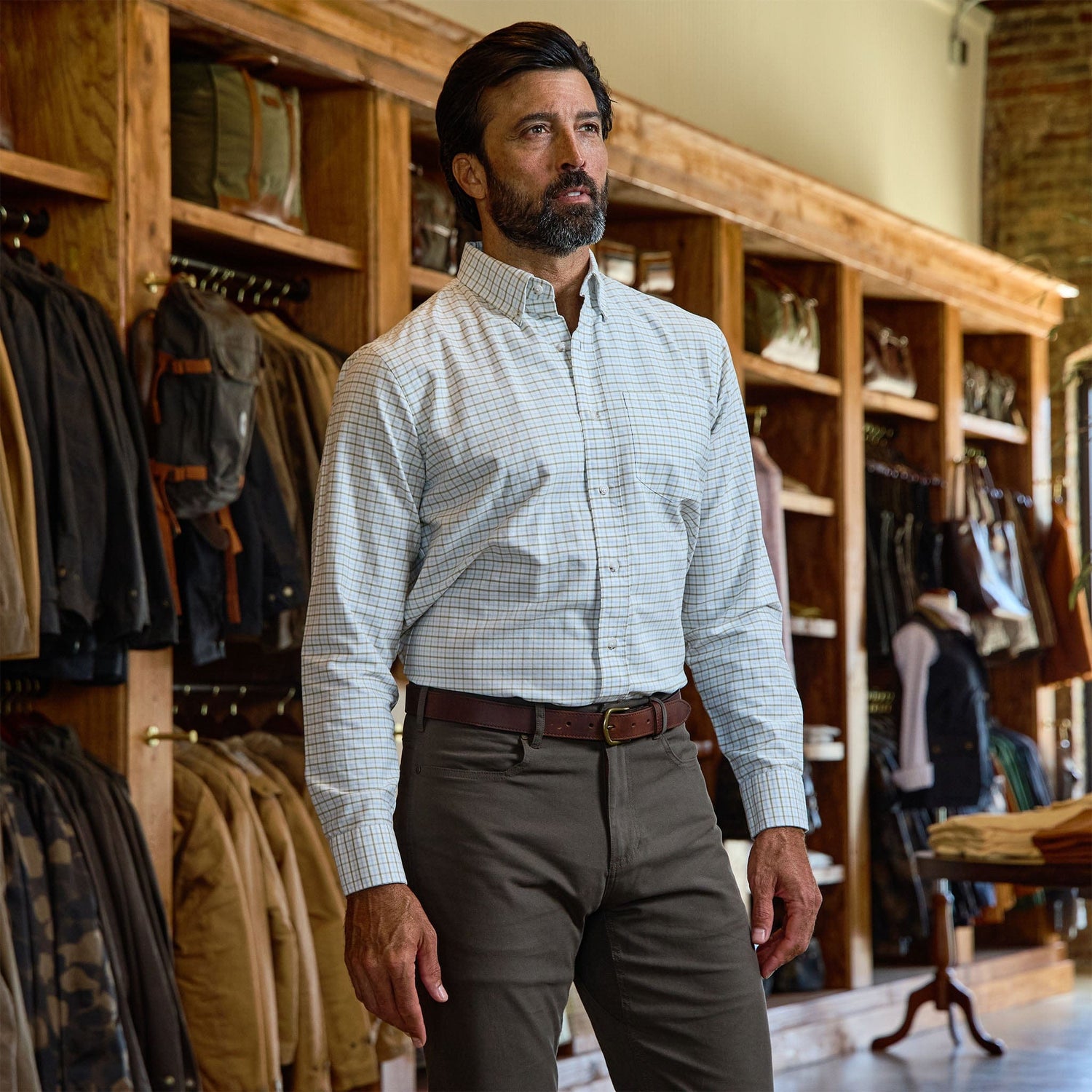 A man in a Tom Beckbe Culver Oxford Shirt and khaki pants stands in a store with wooden shelves and hanging jackets.