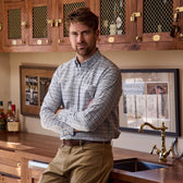 Man in a Tom Beckbe Culver Oxford Shirt stands with arms crossed in a wooden kitchen with framed magazines on the wall.