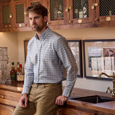 A man in a Tom Beckbe Culver Oxford Shirt and khakis leans against a bar with bottles and framed pictures behind him.
