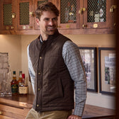 Man in a Tom Beckbe Culver Oxford Shirt stands by a wooden bar with bottles and framed pictures.