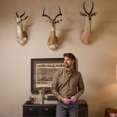 A man in a Tom Beckbe Culver Oxford Shirt stands by a cabinet of bottles and books under three antelope heads.