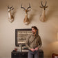 A man in a Tom Beckbe Culver Oxford Shirt stands by a cabinet of bottles and books under three antelope heads.