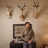 Man in a Culver Oxford Shirt by Tom Beckbe stands by a cabinet with books and bottles, under three antelope heads.