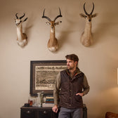 A man in a Tom Beckbe Culver Oxford Shirt stands by a cabinet beneath antelope heads and a framed map.