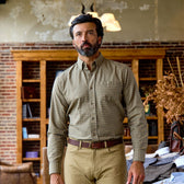 A bearded man in a Tom Beckbe Culver Oxford Shirt stands indoors by a brick wall and wooden shelves.