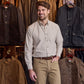 A man wears the Tom Beckbe Culver Oxford Shirt and khaki pants, standing in a store with jackets behind him.