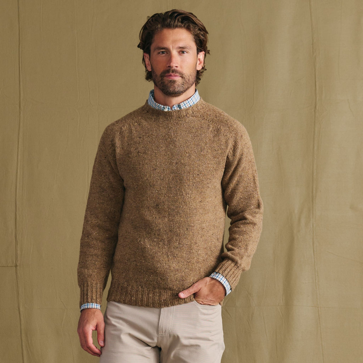 Man in Tom Beckbe Dalton Crewneck Sweater over a blue check shirt and beige pants, set against a tan background.