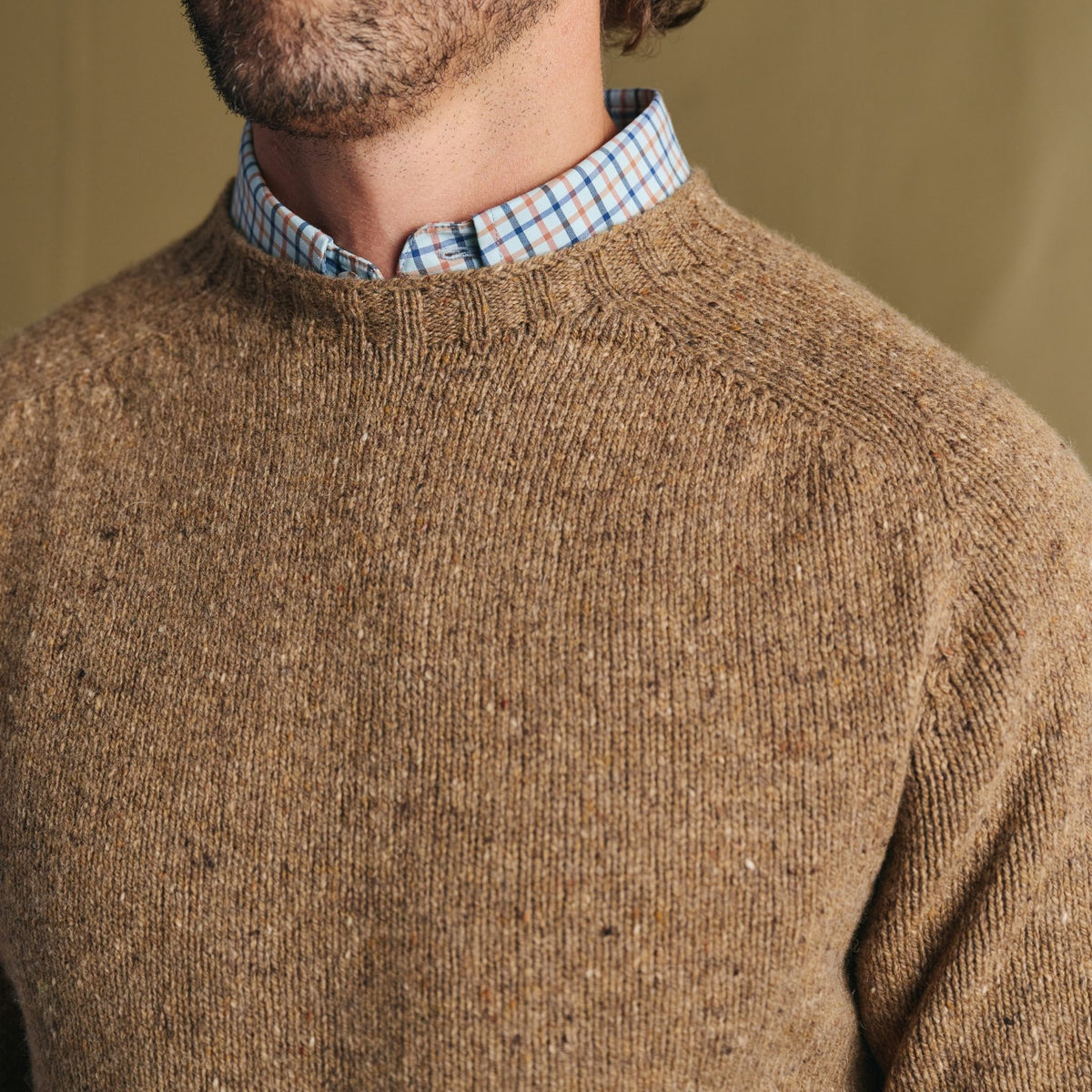 Man wears Tom Beckbe Dalton Crewneck Sweater in brown over a blue and white plaid shirt, shown neck to chest.