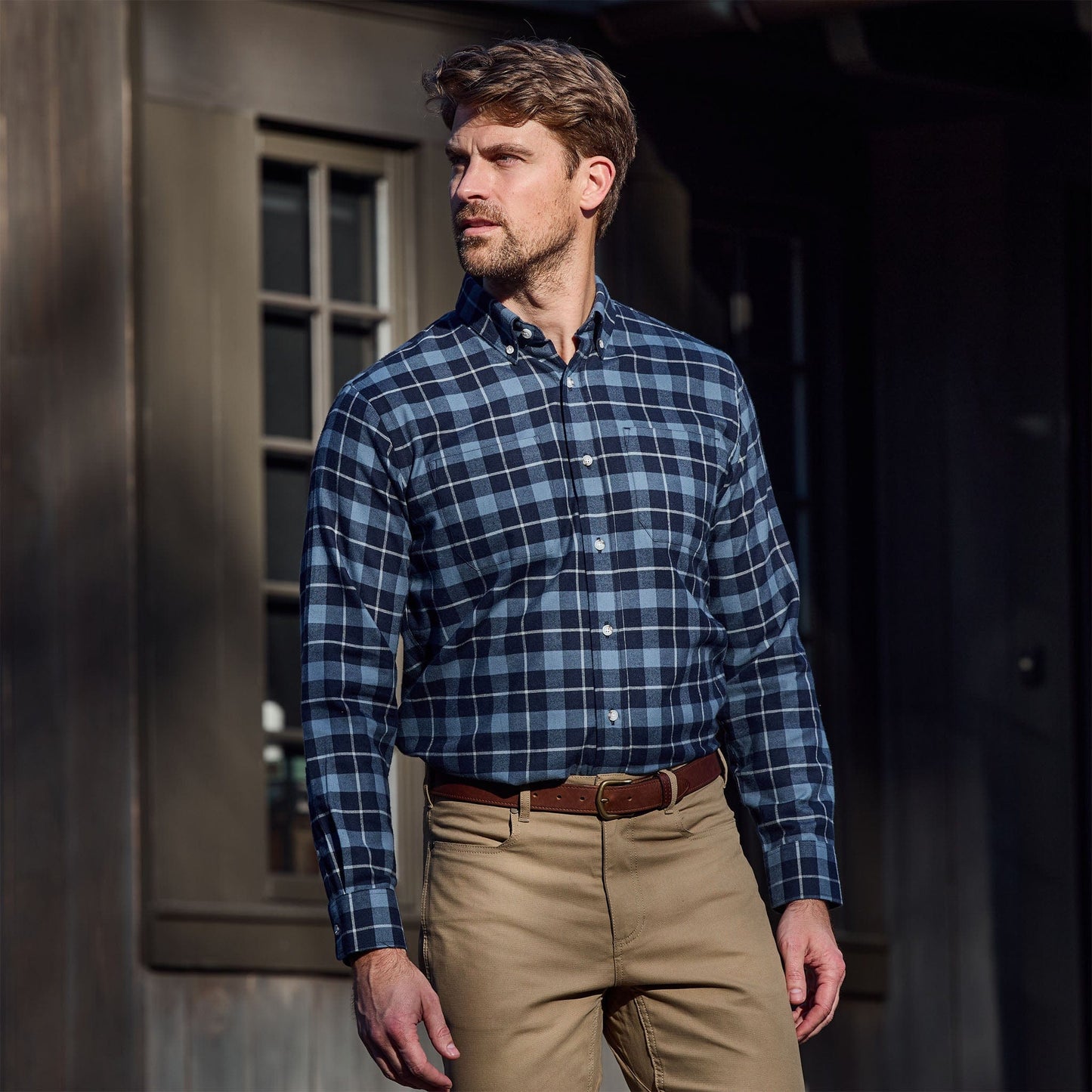 Man wearing a blue plaid button-down shirt and khaki pants standing outside near a building with a window.