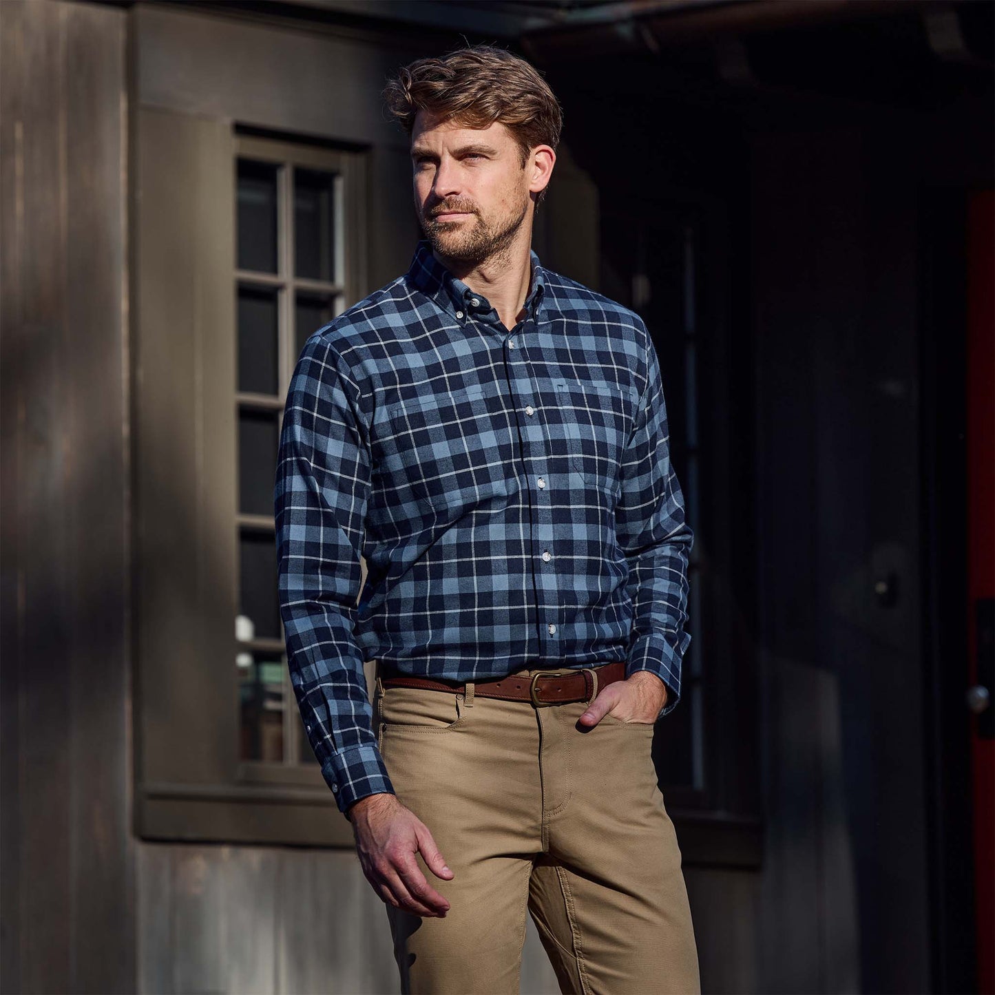 Man wearing a blue plaid shirt and khaki pants stands outside near a window with one hand in his pocket.