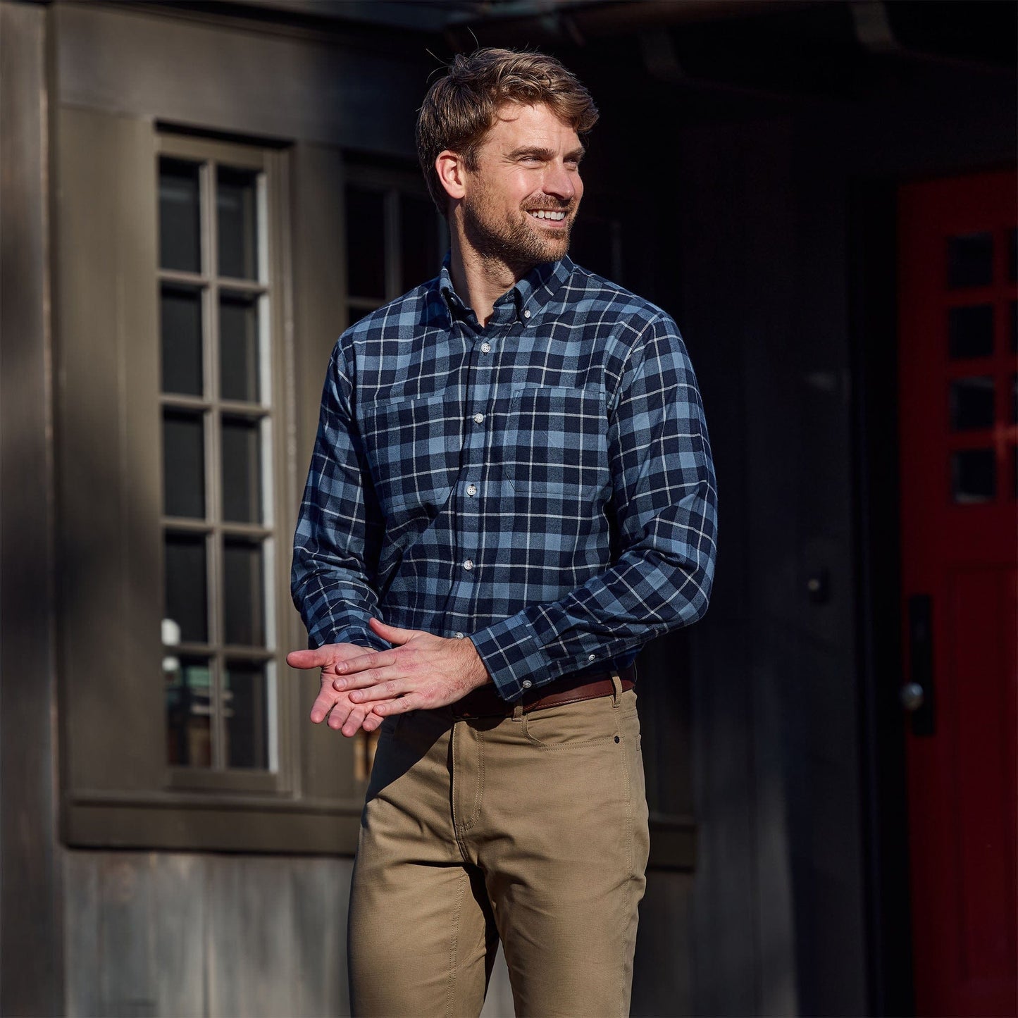Man wearing a blue plaid shirt and khaki pants stands outside near a building with windows and a red door.