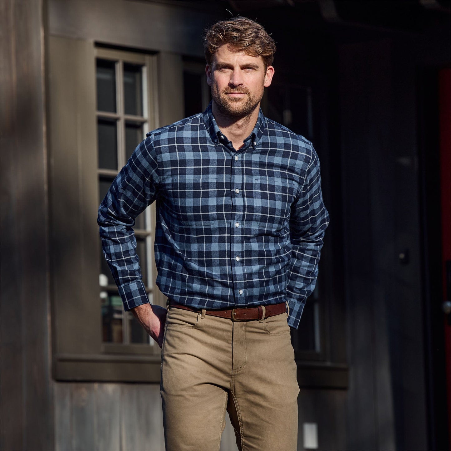 Man wearing a blue plaid shirt and khaki pants stands outside in front of a dark building with windows.
