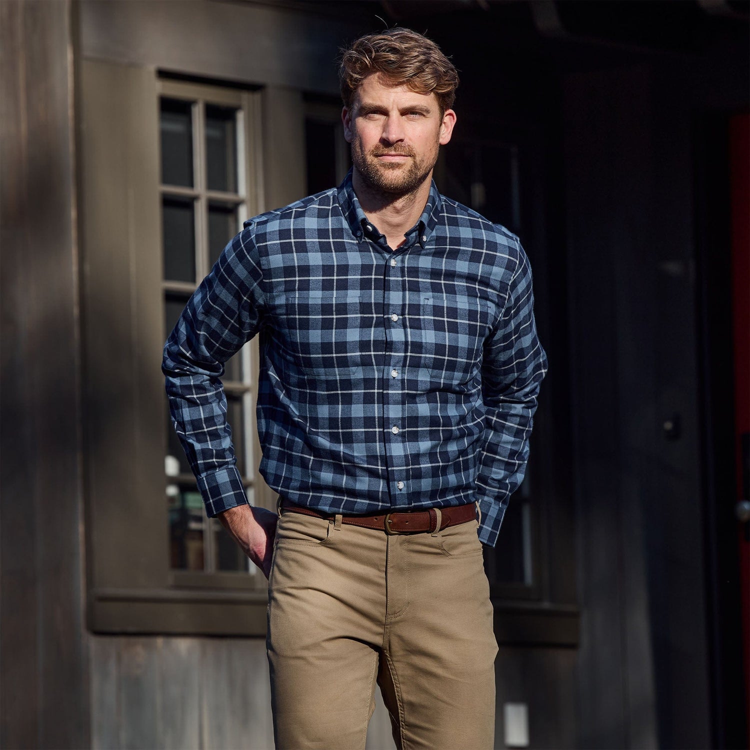 Man wearing a blue plaid shirt and khaki pants stands outside in front of a dark building with windows.