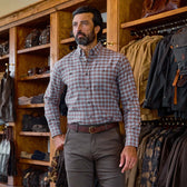 A man wears a Tom Beckbe Dixon Brushed Cotton Twill Shirt and brown pants in a store with wooden shelves behind him.