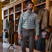 A man in a Tom Beckbe Dixon Brushed Cotton Twill Shirt and gray pants stands among jackets, bags, and clothing in a store.