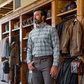 A man wears the Tom Beckbe Dixon Brushed Cotton Twill Shirt and brown pants, surrounded by jackets and bags.