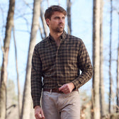 Man wearing Tom Beckbe Dixon Brushed Cotton Twill Shirt in brown plaid and beige pants standing in a sunlit forest.