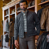 A man in a Tom Beckbe Dixon Brushed Cotton Twill Shirt stands in a clothing store with jackets and bags on shelves.