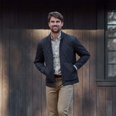 Man in a black quilted jacket, Tom Beckbe Dixon Brushed Cotton Twill Shirt, and khaki pants stands by a dark wood building.