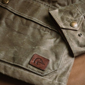 Close-up of Tom Beckbe Womens Steward Jacket: green waxed cloth, brass snaps, and brown leather duck logo patch.