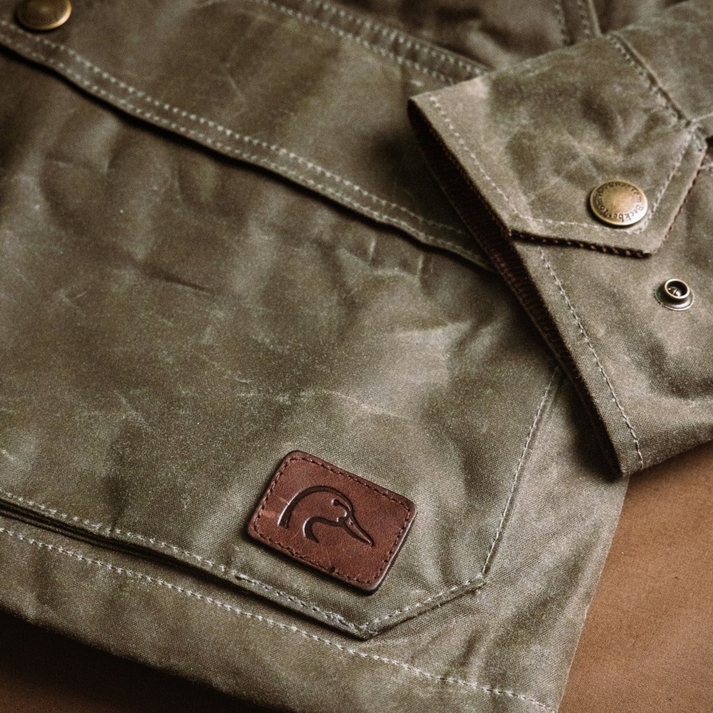 Close-up of Tom Beckbe Womens Steward Jacket: green waxed cloth, brass snaps, and brown leather duck logo patch.