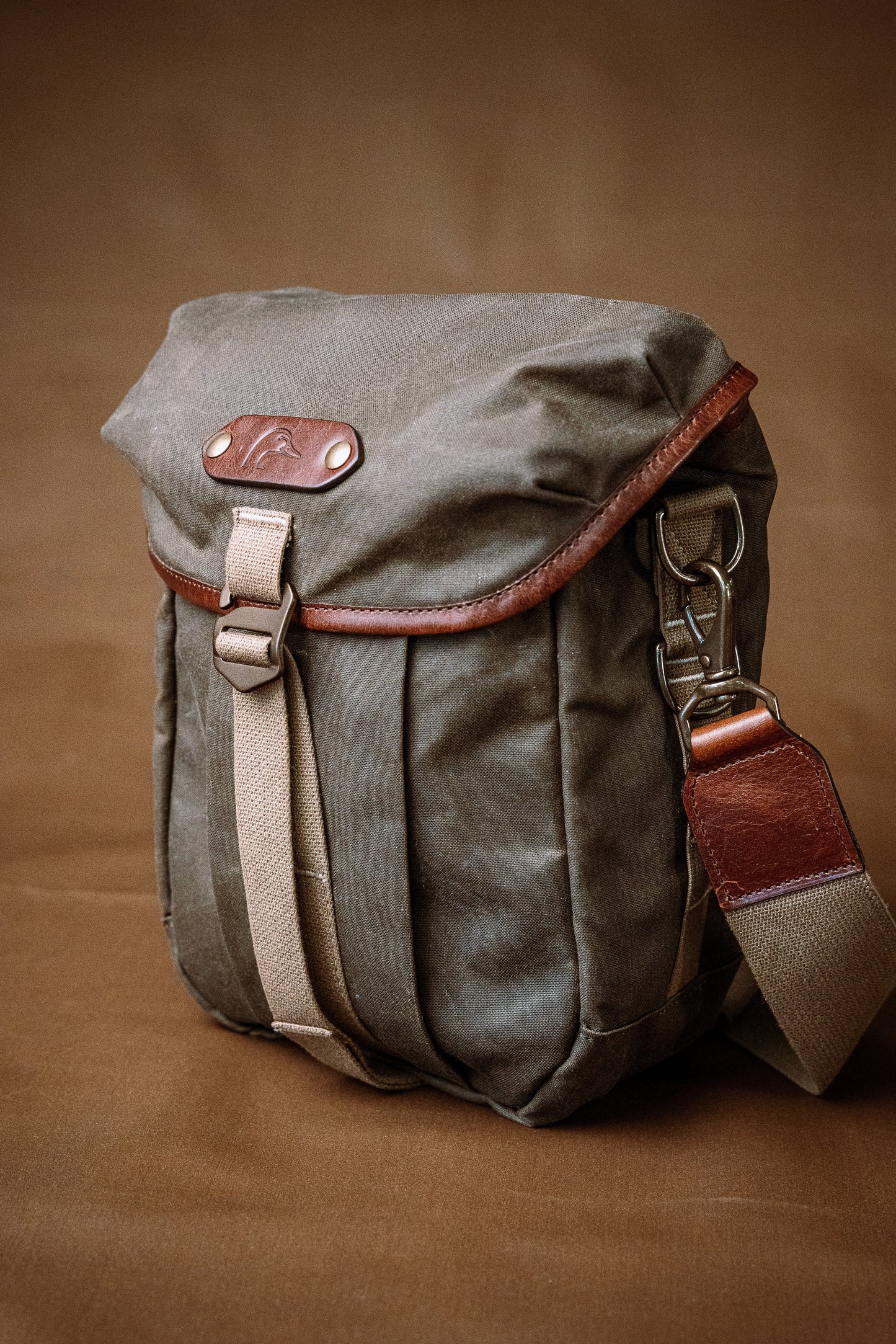The Tom Beckbe DU Timber Bag, waxed cotton canvas with leather strap, sits on a brown background.