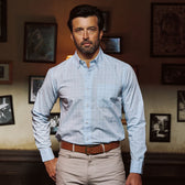 A man with dark hair and a beard wears a Tom Beckbe Ensley Pinpoint Oxford Shirt and beige pants by framed photos.