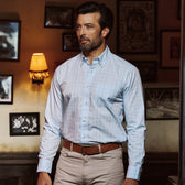 A man wears a Tom Beckbe Ensley Pinpoint Oxford Shirt in light blue checks and beige pants, standing indoors by framed photos.