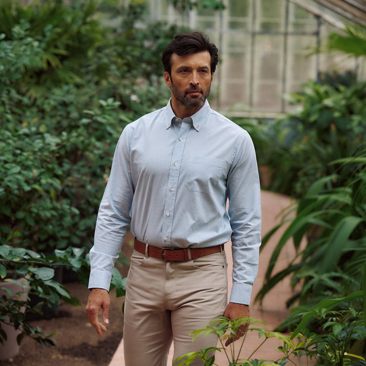 A man in a Tom Beckbe Ensley Pinpoint Oxford Shirt and beige pants walks through a greenhouse of green plants.