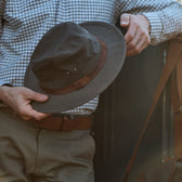 A person in a plaid shirt and brown belt holds a Tom Beckbe Field Hat while leaning on a vehicle.