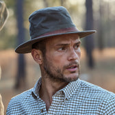A man in a Tom Beckbe Field Hat and plaid shirt looks to the side against a natural, outdoor background.